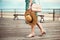 Gorgeous woman wearing summer dress, shoes, pink bag, holding hat and walking on seashore boardwalk at sunset time.