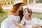 Gorgeous young woman in trendy hat with white ribbon going to kiss daughter in forehead. Laughing dark-haired girl with
