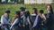 Graduates in black suits eating pizza in a city meadow.