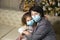Grandmother and granddaughter on the sofa in the living room with Christmas decor hugging in medical masks on their faces. A famil