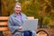 Grandmother sits with a laptop on a bench in an autumn park