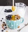 Granola with blueberry, mint, honey and milk in blue bowl with milk jug on a white table and metal spoon