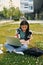 On the grass with laptop on legs. Young asian woman is outdoors at daytime