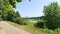 On a grassy hill in the city park, there is a tiled walkway among the trees. From above, there is a view of the river with its ban