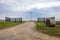 A gravel driveway leads past a round hay bale through an open gate up a gently rolling hill to the farm beyond