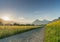 Gravel road parting a rapeseed canola field and a yellow wildflower meadow with the setting sun disappearing behind a beautiful mo