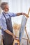 gray bearded artist painting on easel, canvas