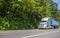 Gray big rig semi truck transporting cargo in covered bulk semi trailer driving on the road with trees on the road side