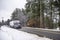 Gray big rig semi truck transporting cargo in refrigerator semi trailer running on the local winter road with snow and trees on