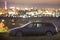 Gray car parked at night in green meadows on background of lights of distant city buildings and dark mountain ridge under bright