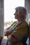 A gray-haired elderly woman stands at the window with her arms crossed over her chest