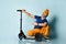 Gray-haired grandpa in t-shirt, sunglasses, orange pants, hat, gumshoes. Holding scooter, squatting on blue background. Close up