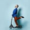 Gray-haired male in white shirt, jacket, brown pants and loafers. Riding black scooter and posing sideways on blue background