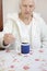 Gray-haired old man sits at the table and takes a cup of tea from a teaspoon.