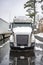 Gray industrial big rig semi truck with semi trailer standing on the winter truck stop parking lot with snow and ice on the hood