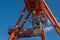 Gray mobile cab with lifting hook, located on rail of huge orange overhead crane, electric shields are mounted on left.