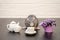 Gray rat sits with cup of tea and teapot, with bouquet of purple flowers on black and white wooden background