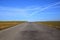 A gray road with spotty pavement stretches straight to the horizon with fields in winter that are brown and empty against a blue