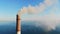 Gray smoke comes out of a large brick chimney in the factory against a clear blue sky and a large lake.