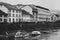 Grayscale shot of the boats on the river against the buildings on the shore.