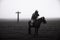Grayscale shot of a male in rain clothes riding a horse in a field with a cross in the background