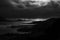 Grayscale shot of silhouettes of hills and a lake under the sky with dark clouds