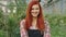 Great looking with large smile redhead gardener woman closeup to the camera in a flower greenhouse looking to the camera