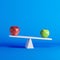 Green apple sitting on seesaw with red apple on opposite end on blue background