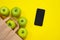 Green apples rolling out from the cardboard package on bright yellow surface next to smart phone. Craft organic pack