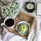 Green and blue hydrangea branch, a large cup of tea, a donut on a saucer, a tea strainer, an apple, on a tray, an alarm clock, a