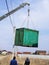 Green cargo container which gray boom with blue hook of truck manipulator lift up near the power line.