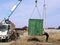 Green cargo container which gray boom with blue hook of truck manipulator put on the ground near the power line.