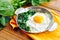 Green cooked spinach with fried egg on iron pan plate. Fried eggs healthy breakfast on wooden table background on orange napkin