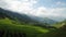 Green fields with firs, snowy mountains and large clouds. A clear blue sky.