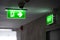 A green fire exit sign is placed on the ceiling along the dimly lit corridor and there is green exit sign on the exit door