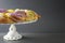 A green, gold, and purple festive Mardi Gras King Cake rests on a white cake stand with a traditional baby figurine on top with a