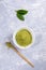 Green japanese matcha tea powder in white bowl, special wooden bamboo spoon and fresh leaves on grey textured backdrop.