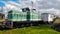 Green locomotive parked close to the main train station with small white railway carriage being connected to it