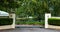 Green metal wrought iron driveway property entrance gates set in concrete  fence, garden shrubs, trees, lights