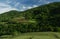 Green mountainsides with trees and a vegetable garden under a blue cloudy sky.