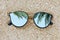 Green palm leaves mirroring in sunglasses on sandy beach, top view
