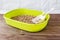 Green plastic cat litter box with wooden pellets and scoop on a brown wooden floor. New cat toilet tray near the wall. Toilet for