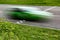 A green race car at speed crosses the finish lane of an asphalt track against a background of green grass and bushes