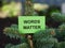 A green sticky note with the phrase Words Matter on it being held up by a fir tree branch