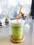 Green tea milk frappe in clear plastic glass have orange straw in wooden tray put on dining table with cactus, succulent and mirro