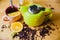 Green teapot on a wooden table. Next to a slice of orange, full cup of tea, honey jar and scattered tea leaves and herbs