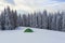 Green tent stands on the snowy lawn in the cold winter day. High spruce trees. Touristic camping rest place.