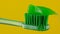 Green toothpaste is placed on a green toothbrush on a yellow background in close-up.