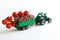 Green toy tractor is carrying a branch of fresh red cherry tomatoes in a trailer. White background. The concept of agricultural