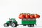 Green toy tractor is transporting a branch of fresh red cherry tomatoes in a trailer. White background. Harvest and delivery